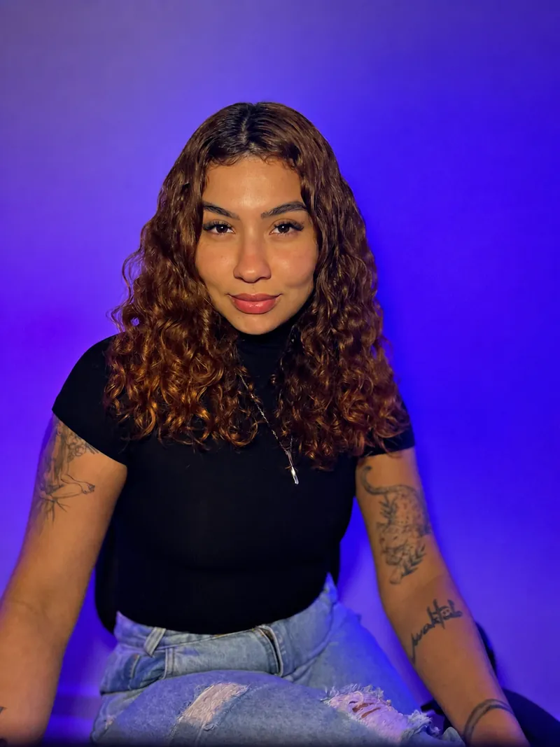Victoria sentada em estúdio com fundo roxo, sorrindo pra câmera.